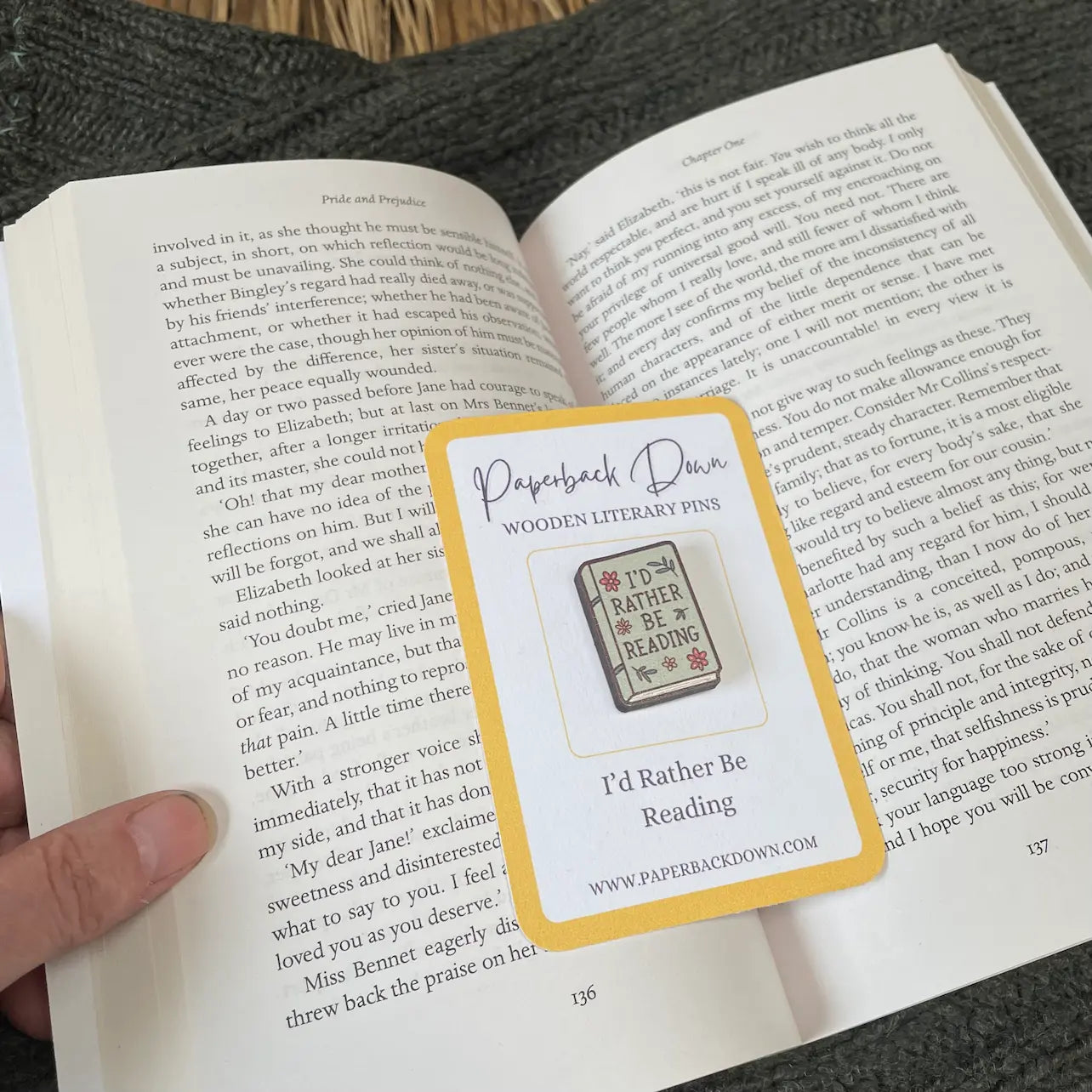 Wooden literary pin labeled 'I'd Rather Be Reading' on a book page with a card from 'Paperback Dawn'.