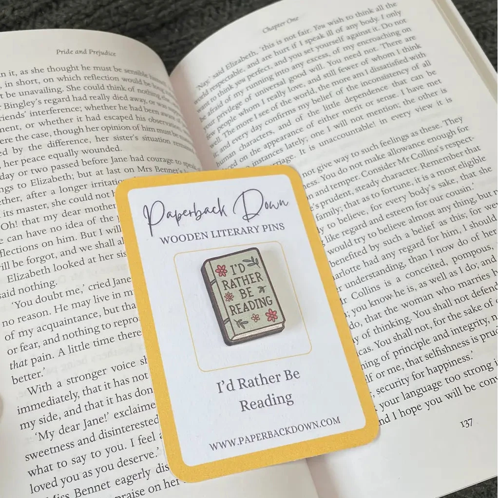 Wooden literary pin labeled 'I'd Rather Be Reading' on a book page with a card from 'Paperback Dawn'.