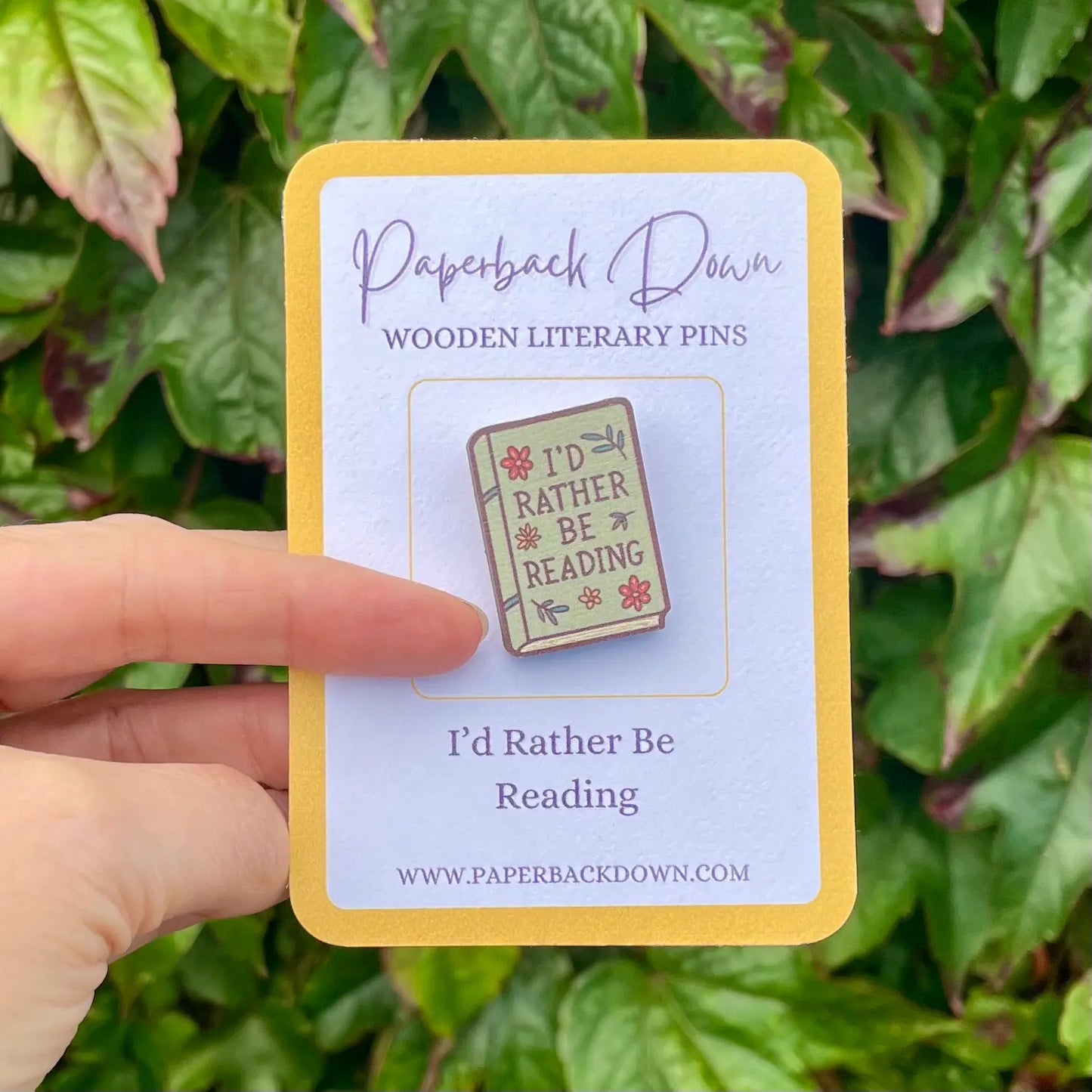 Pin with 'I'd Rather Be Reading' design on a card held by a hand against a leafy background