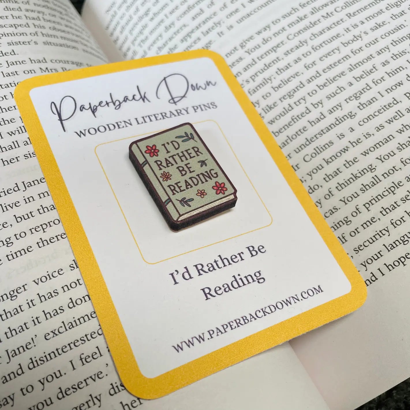 Enamel pin with 'I'd Rather Be Reading' design on a card, placed on an open book.