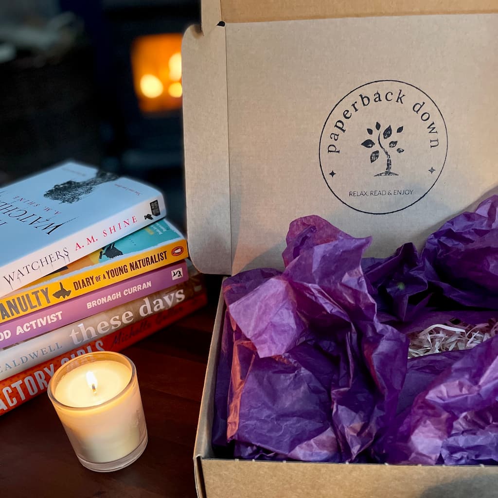 An open box with the Paperback Down logo on the lid, hinting at the subscription box contents inside. A stack of books by Irish authors is beside the box and a lit candle in front of a cosy fireplace