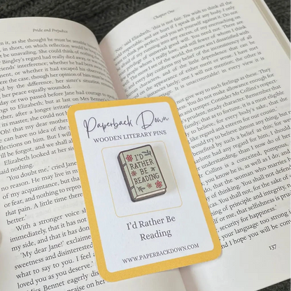 Wooden literary pin labeled 'I'd Rather Be Reading' on a book page with a card from 'Paperback Dawn'.