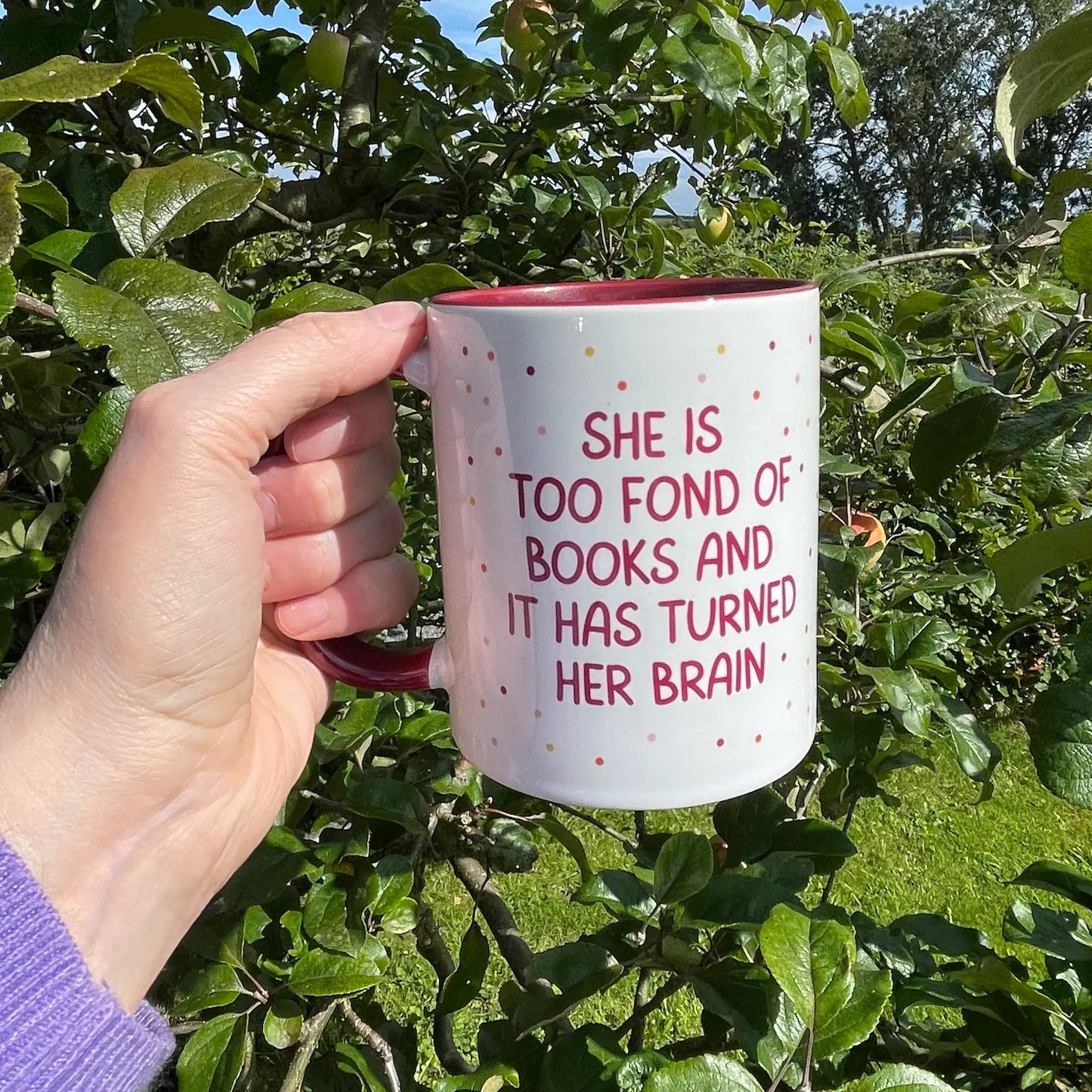 Image shows a hand holding a mug in front of a tree - the mug shown is a Book  Mug with the Little Women  quote- She is too fond of books and it has turned her brain - Red and White Ceramic Mug with Polka Dot pattern