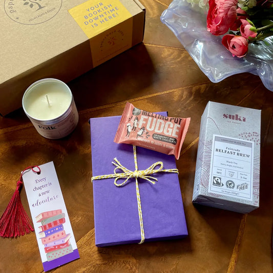 Image shows a gift box for those who love tea and books. Pictured is a beautifully wrapped book, a scented Field Day Candle with notes of tea leaves and nutmeg, locally made fudge, a box of Suki Belfast Brew Tea and a tassel bookmark.