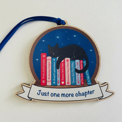 Decorative ornament with a black cat and books design, featuring the text 'Just one more chapter'.